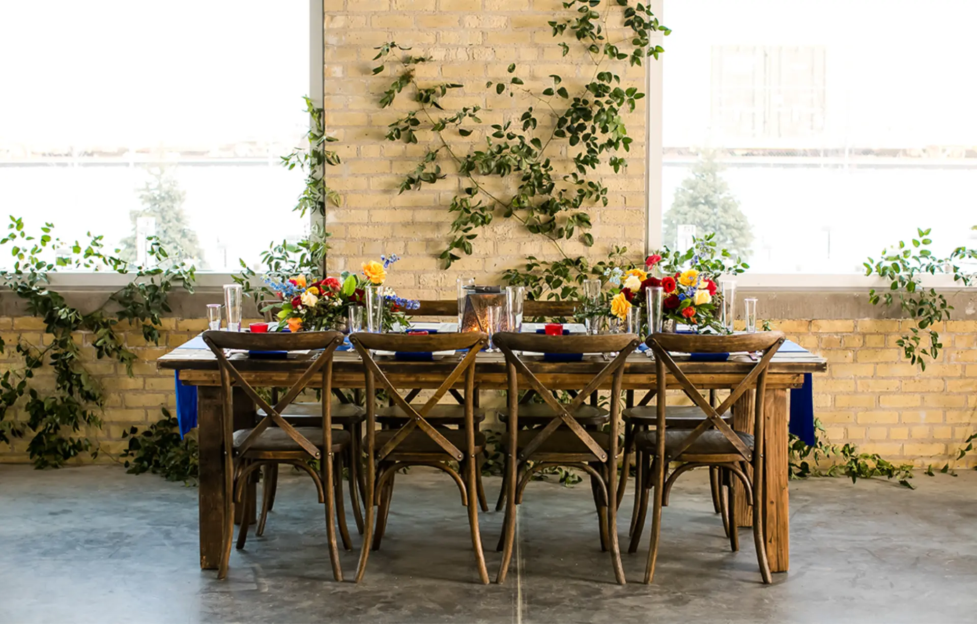A long wooden table with bright florals in front of a brick wall and tall windows